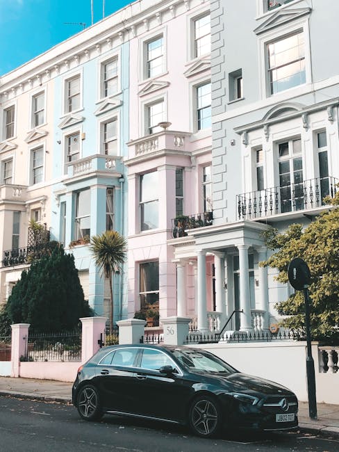 A black hatchback car parked on a residential street in front of a row of Georgian-style terraced houses with ornate white facades, large sash windows, and small balconies with wrought iron railings, in the Westbourne Grove area of Notting Hill. The pavement is lined with a small palm tree, shrubs, and a decorative fence surrounding garden areas. The scene is well-lit with natural daylight, and the street appears clear of traffic. This setting illustrates a typical scene for home relocation or furniture transport, with potential access points for moving teams such as Man and a Van Notting Hill, involved in professional removals and packing services. The overall environment emphasizes the importance of understanding parking options and timing for efficient moving or house clearance, especially in a sought-after area like Notting Hill.