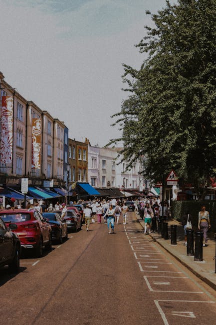 Photograph of a residential street featuring a row of white, multi-storey Victorian-style terraced houses with ornate detailing and bay windows. The buildings are illuminated by natural daylight, casting soft shadows on their facades. Several parked cars, including sedans and hatchbacks, line the pavement in front of the houses, with some partially visible to the left and right sides of the image. The street is calm and free of moving vehicles, with a large tree visible to the left, its branches extending above the rooftops. The area appears quiet, with no pedestrians or movers present, highlighting a typical urban setting suitable for home relocation activities. The scene conveys a clean, orderly environment conducive to professional removals services, such as loading furniture and packed boxes into a van, with [COMPANY_NAME] possibly preparing for a move along Westbourne Grove, NOTTING HILL, aligning with the theme of packing and furniture transport during a house removal.