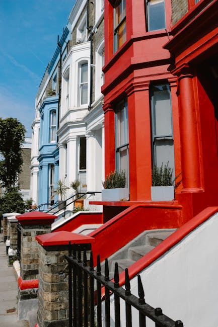 A row of colorful terraced houses on a busy street in Notting Hill during daytime, with a man in a dark jacket and a woman in a red coat carrying a cardboard box wrapped in plastic moving blankets, pa