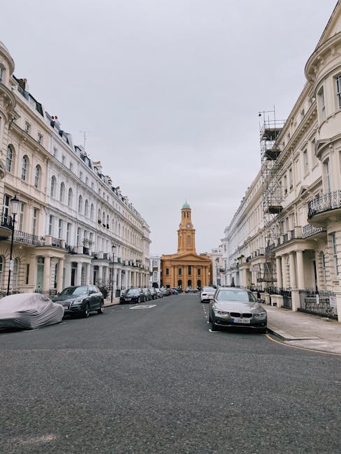 A wide view of a residential street in Notting Hill lined with white Victorian-style terraced houses featuring black wrought iron balconies and large sash windows. Parked cars, mostly covered with protective coverings or parked directly on the pavement, line both sides of the street. In the distance, a historic church with a tall, square tower topped by a small dome and clock faces is visible, set against a cloudy sky. The scene depicts an urban environment suitable for house removals and furniture transport, with the street prepared for an ongoing moving or packing process. Man and a Van Notting Hill's removals service may include loading household furniture and boxes from this location, with equipment such as trolleys and straps potentially in use to facilitate home relocation and loading onto their vehicle for transport.