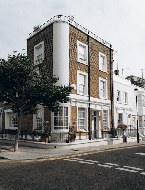 A wide view of a residential street in Notting Hill lined with white Victorian-style terraced houses featuring black wrought iron balconies and large sash windows. Parked cars, mostly covered with protective coverings or parked directly on the pavement, line both sides of the street. In the distance, a historic church with a tall, square tower topped by a small dome and clock faces is visible, set against a cloudy sky. The scene depicts an urban environment suitable for house removals and furniture transport, with the street prepared for an ongoing moving or packing process. Man and a Van Notting Hill's removals service may include loading household furniture and boxes from this location, with equipment such as trolleys and straps potentially in use to facilitate home relocation and loading onto their vehicle for transport.